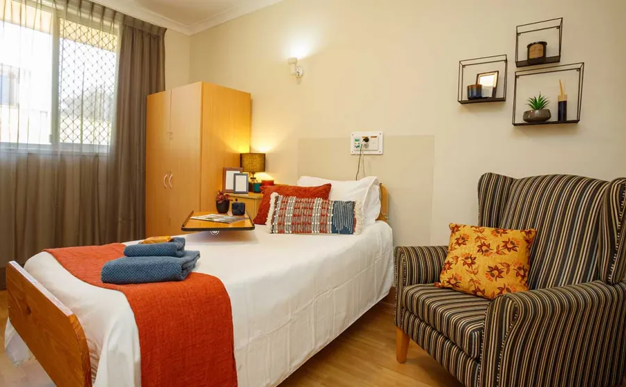 A bedroom with a single bed and striped armchair at Caloundra Place Care Community.